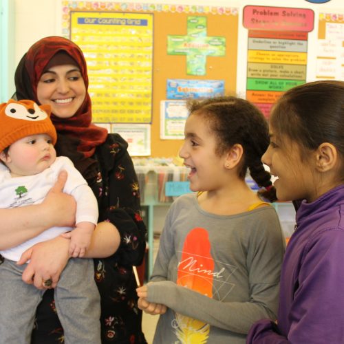 Two girls sing to baby while Mom looks on smiling